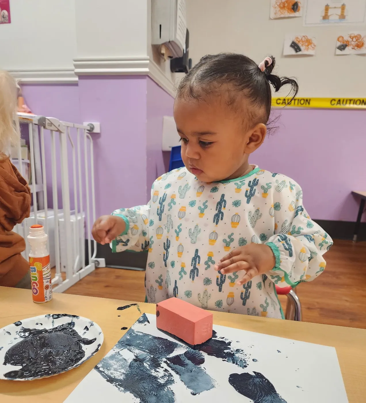 A toddler in a cactus-print smock stamping paint onto paper